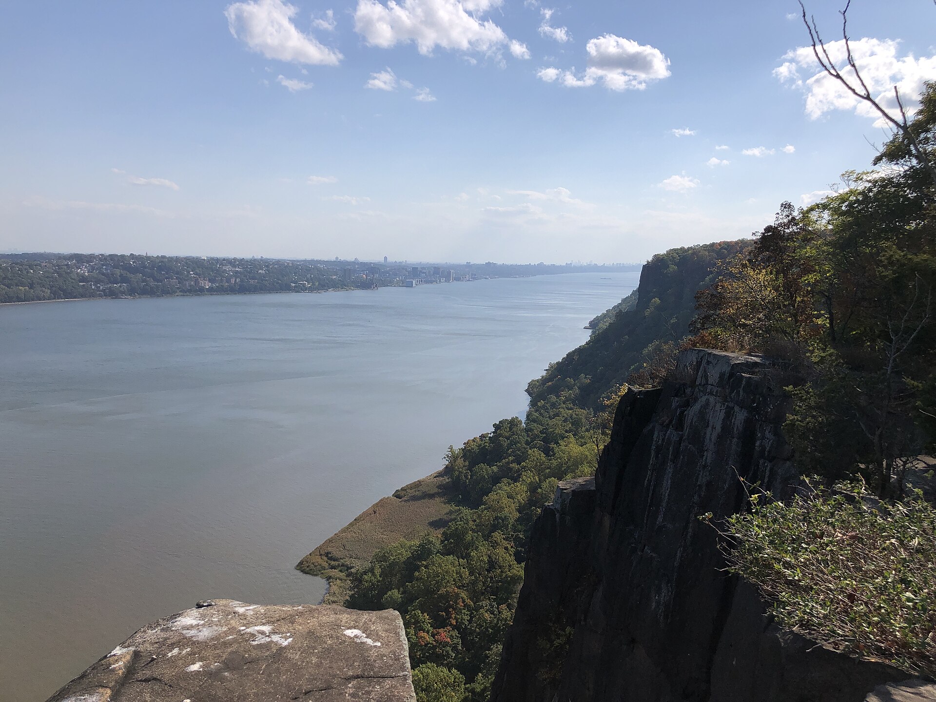 The Hudson River seen from the top of the Palisades cliffs, with a sheer dark rock face on the right and towns on the New York shore in the distance