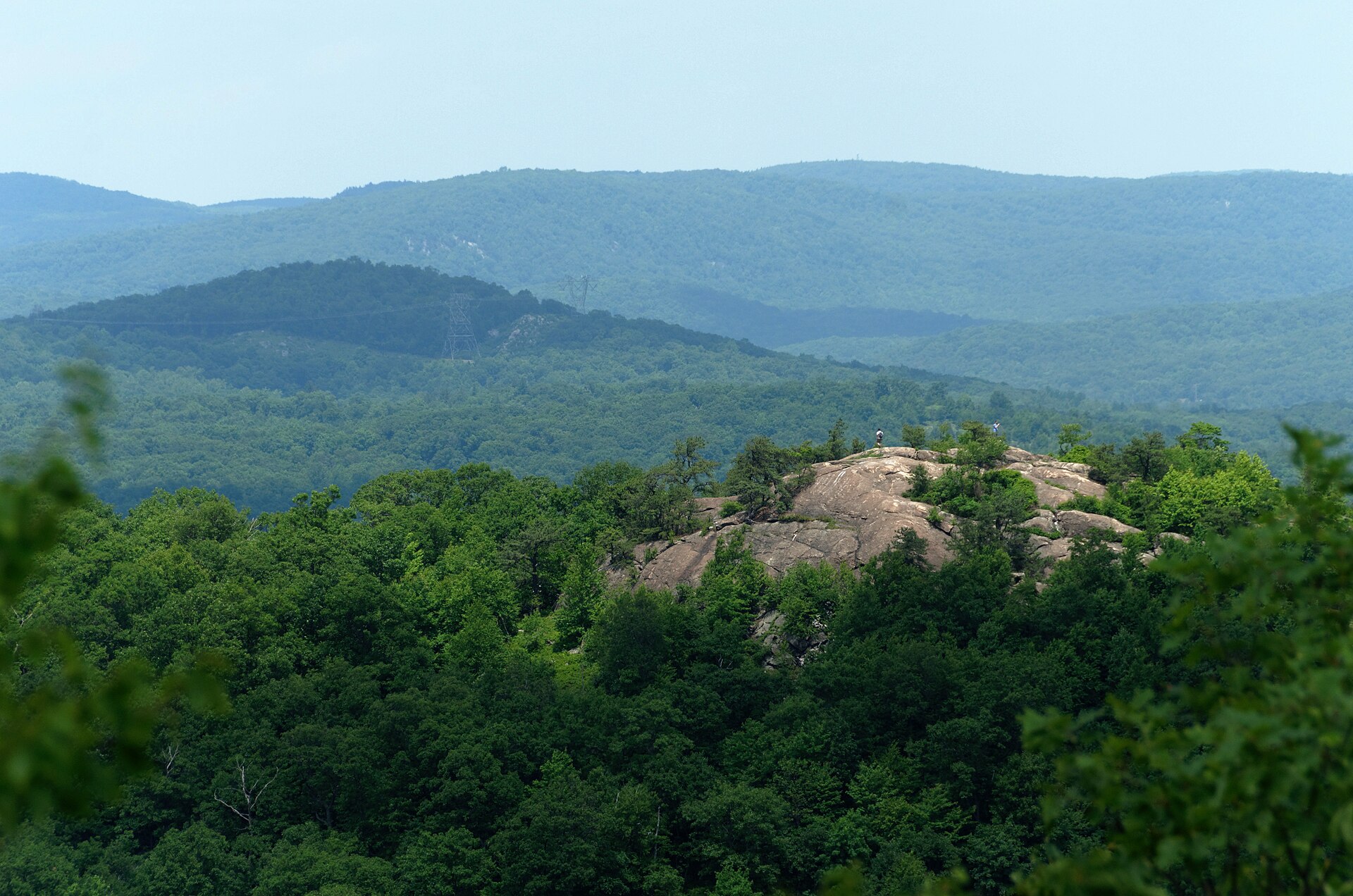 The bare rock dome of Wyanokie High Point rising out of dense green forest, with two tiny figures standing on the summit and wooded ridges behind