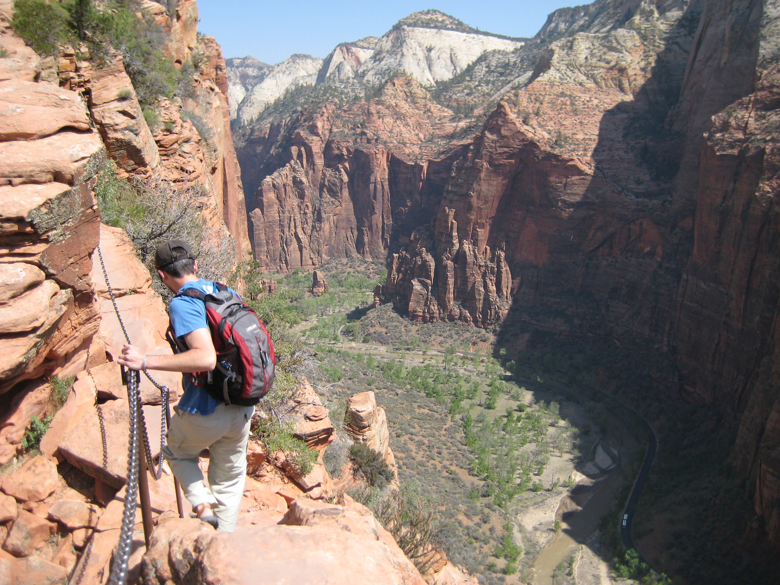 A hiker holding a chain bolted to the rock on the Angels Landing spine, looking down more than a thousand feet into Zion Canyon