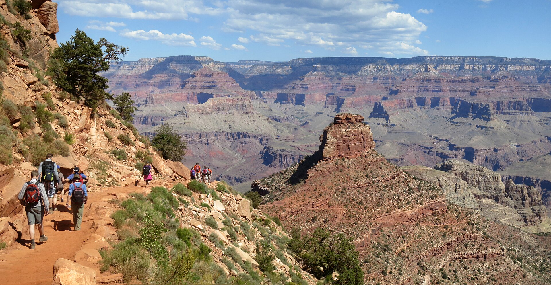 A line of hikers descending a red dirt trail cut into the wall of the Grand Canyon, with buttes and layered cliffs stretching to the horizon
